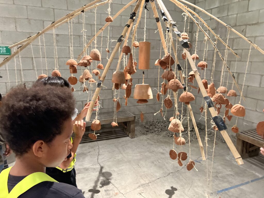 A photo of some students looking at a hanging art piece consisting of fired clay pieces on strings suspended from wood. It looks like it would make noise in a breeze
